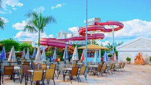 Een waterpark met een grote rode waterglijbaan, buitenzitplaatsen met parasols en een gebouw met de tekst 'Wilson Pedro'.