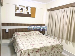 A bedroom with a bed covered in floral bedding, a wooden headboard, a wall-mounted artwork, and a window with curtains.