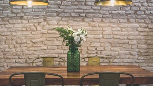 Un tavolo da pranzo con un vaso di fiori verdi, due sedie e due lampade a sospensione, sullo sfondo di un muro in pietra.