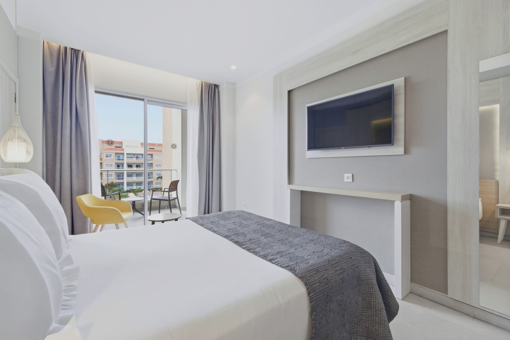 A hotel room with a bed, a TV mounted on the wall, a balcony with chairs, and a view of buildings outside.