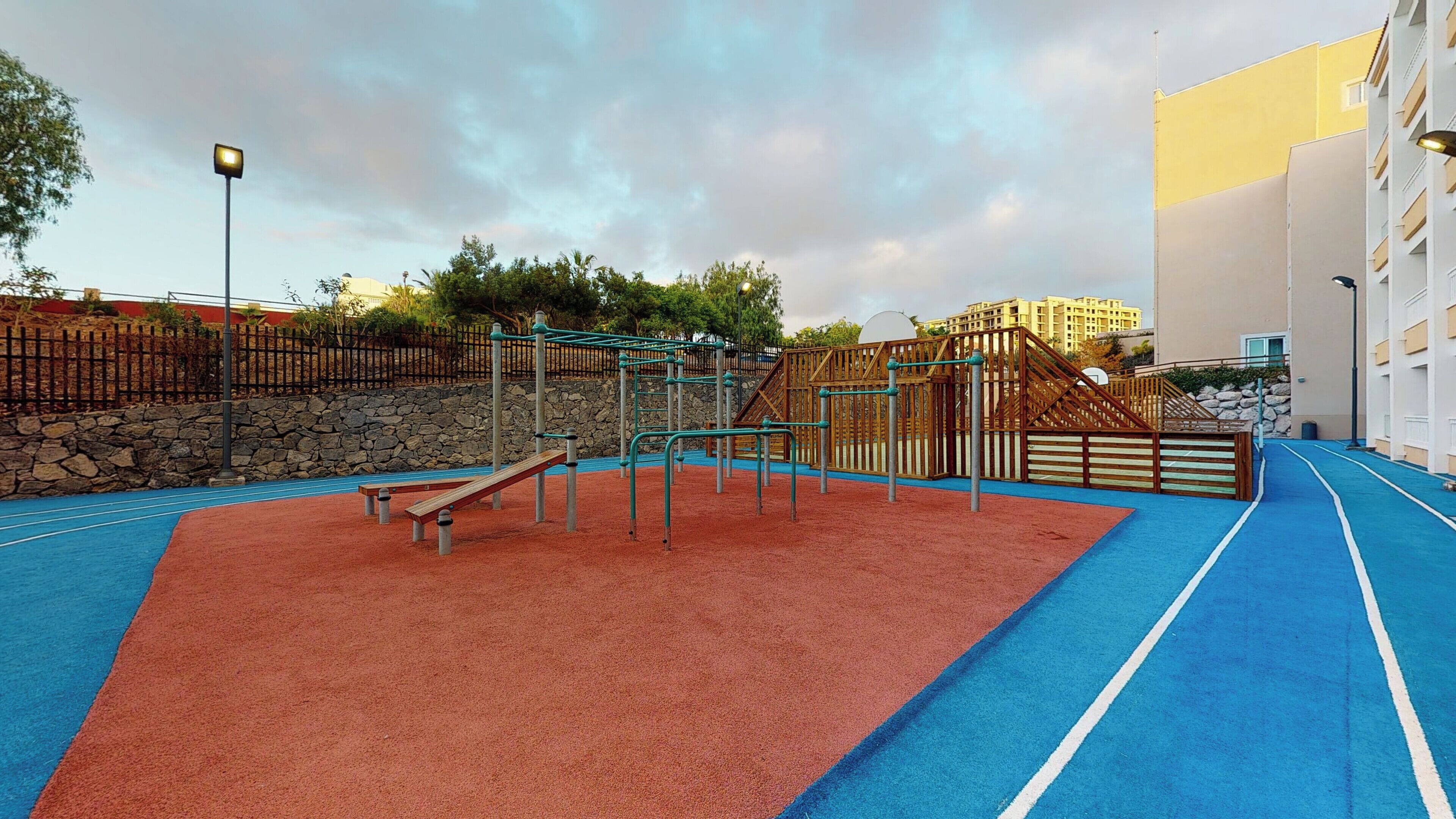 An outdoor playground with a blue and red rubberized surface, surrounded by a stone wall and wooden fencing.