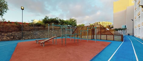 An outdoor playground with a blue and red rubberized surface, surrounded by a stone wall and wooden fencing.