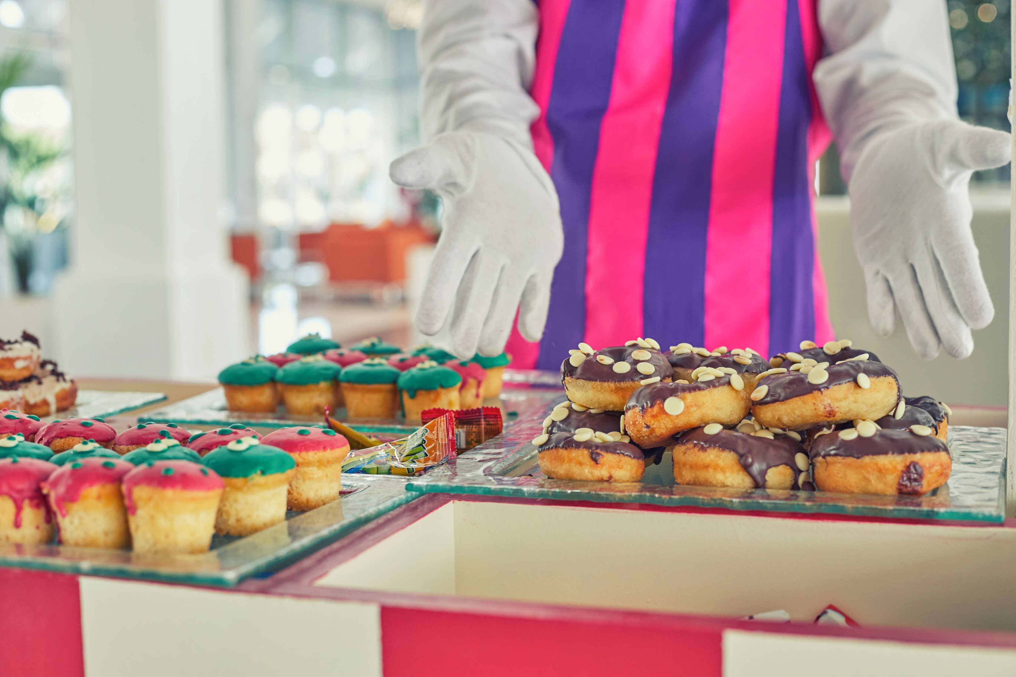 Mostrador de pastelería con varios productos horneados y una persona con mandil y guantes a rayas.