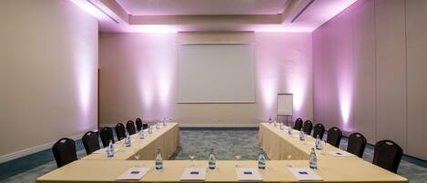 Un salón de conferencias con una larga mesa, sillas, botellas de agua y una pantalla de proyector.