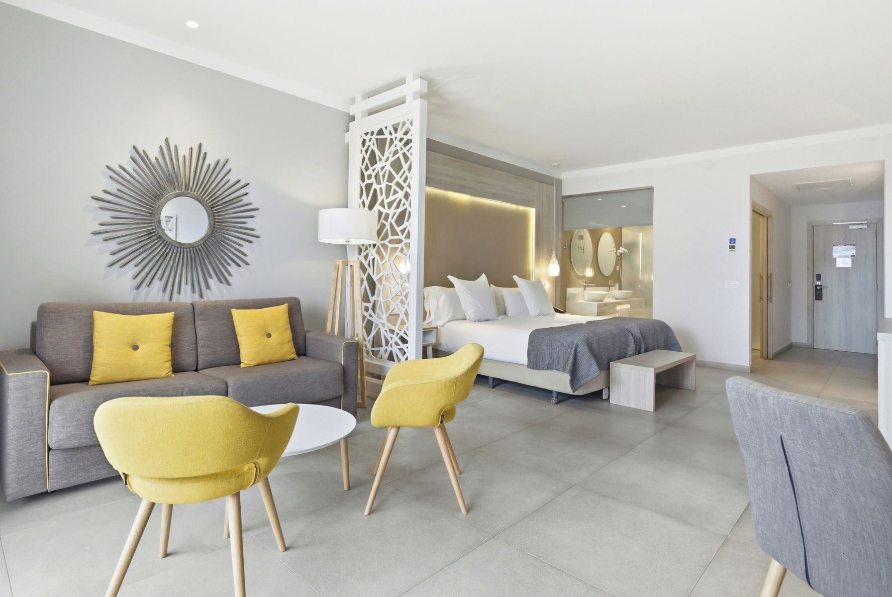 Une chambre d’hôtel moderne avec un canapé, deux fauteuils jaunes, un lit et une salle de bain équipée d’un miroir.
