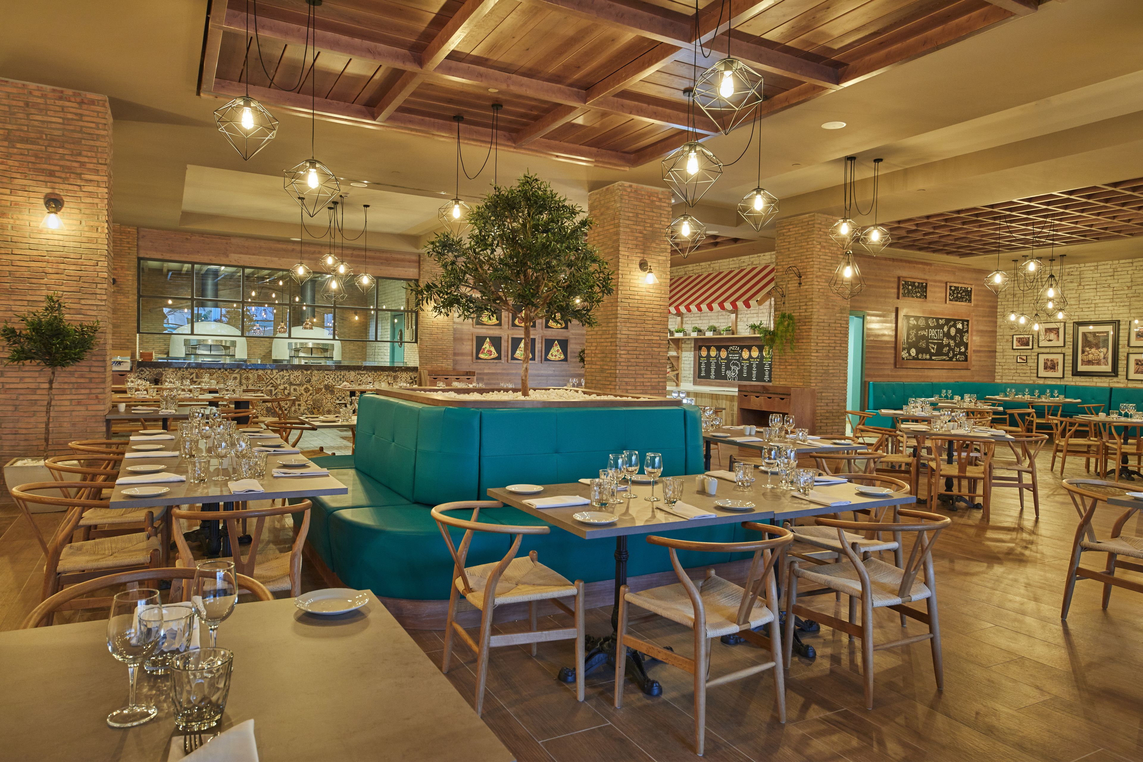 Ein Restaurant mit Holztischen, grün gepolsterten Sitzplätzen, hängenden Lampen und einer Backsteinwand.