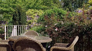 Korbmöbel mit Glastisch und Stühlen, arrangiert in einem Garten mit Metallzaun und unterschiedlichen Pflanzen.