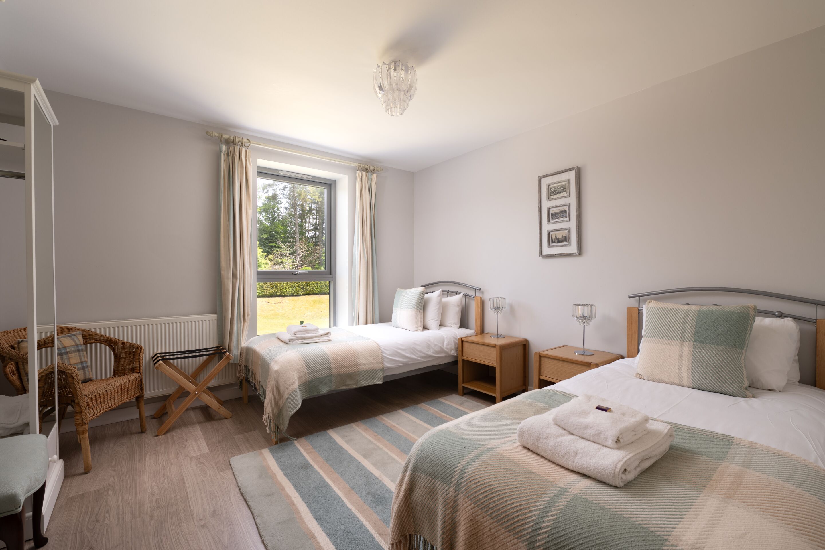 A bedroom with two beds, a window with curtains, a wicker chair, and a bedside table with a lamp.