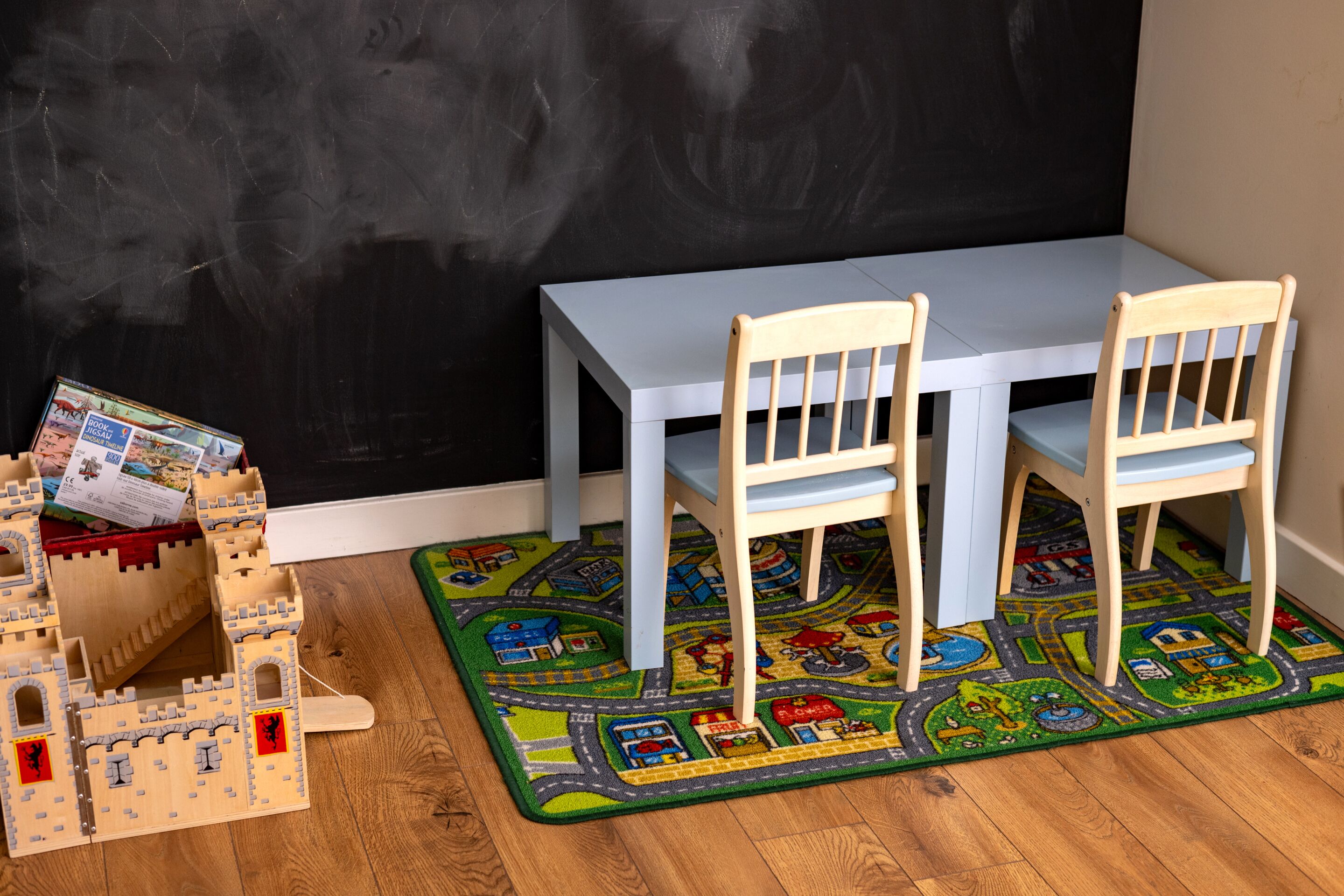 A room with a white table and two wooden chairs, a castle playset, and a colorful play mat.