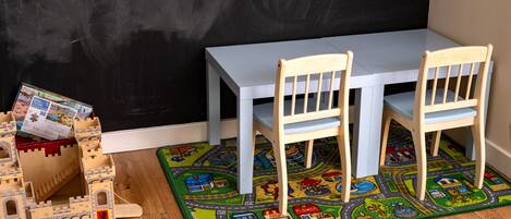 A room with a white table and two wooden chairs, a castle playset, and a colorful play mat.