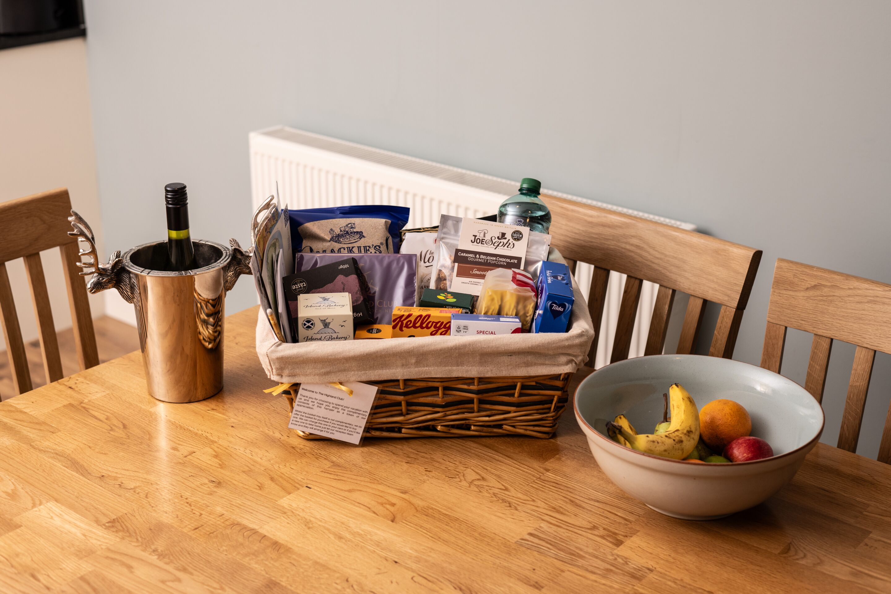 A wooden table with a wine bottle, a basket of snacks, and a bowl of fruit.