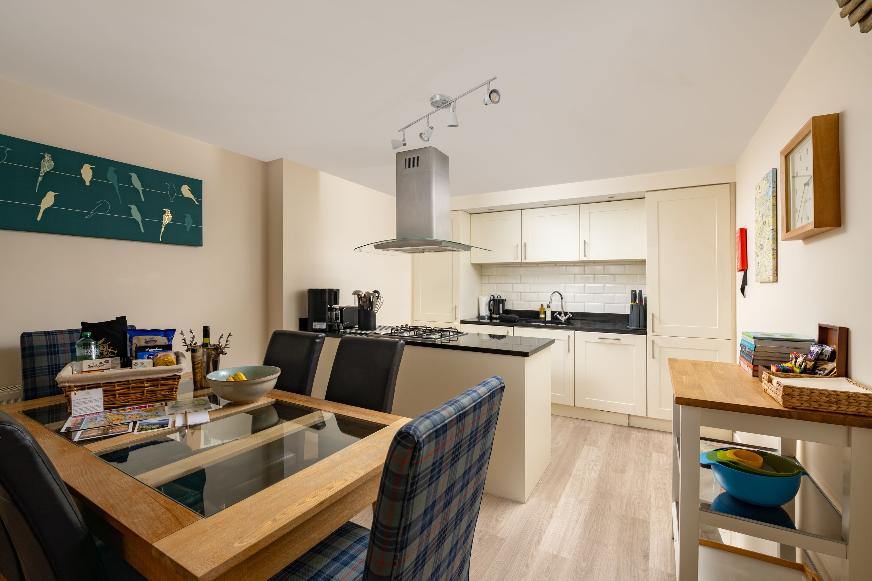 A modern kitchen with a central island, white cabinets, and a dining area with a glass table and plaid chairs.