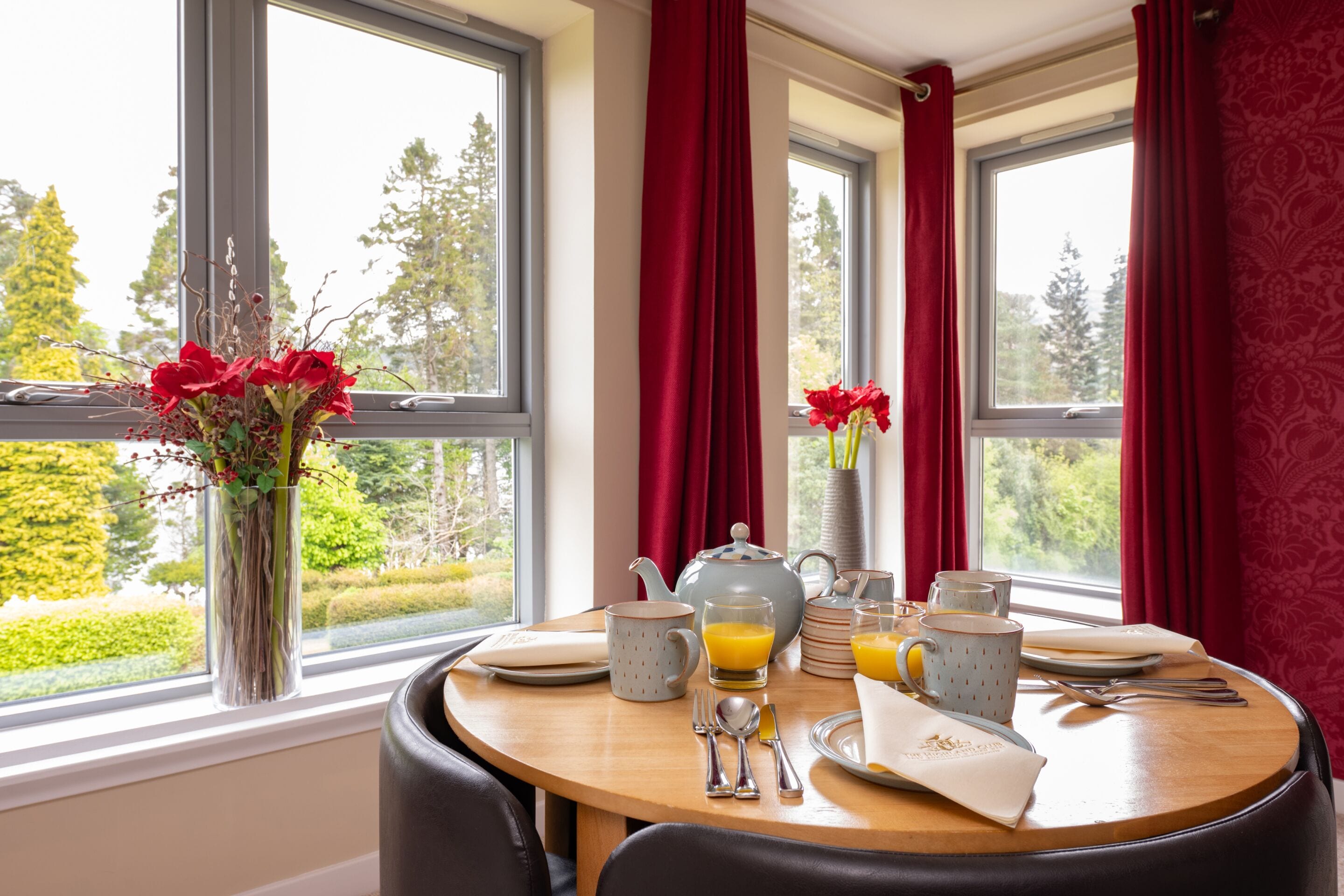 Una mesa de comedor dispuesta con alimentos para el desayuno, un jarrón con flores rojas y vistas a la vegetación a través de amplios ventanales.