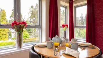 Una mesa de comedor dispuesta con alimentos para el desayuno, un jarrón con flores rojas y vistas a la vegetación a través de amplios ventanales.