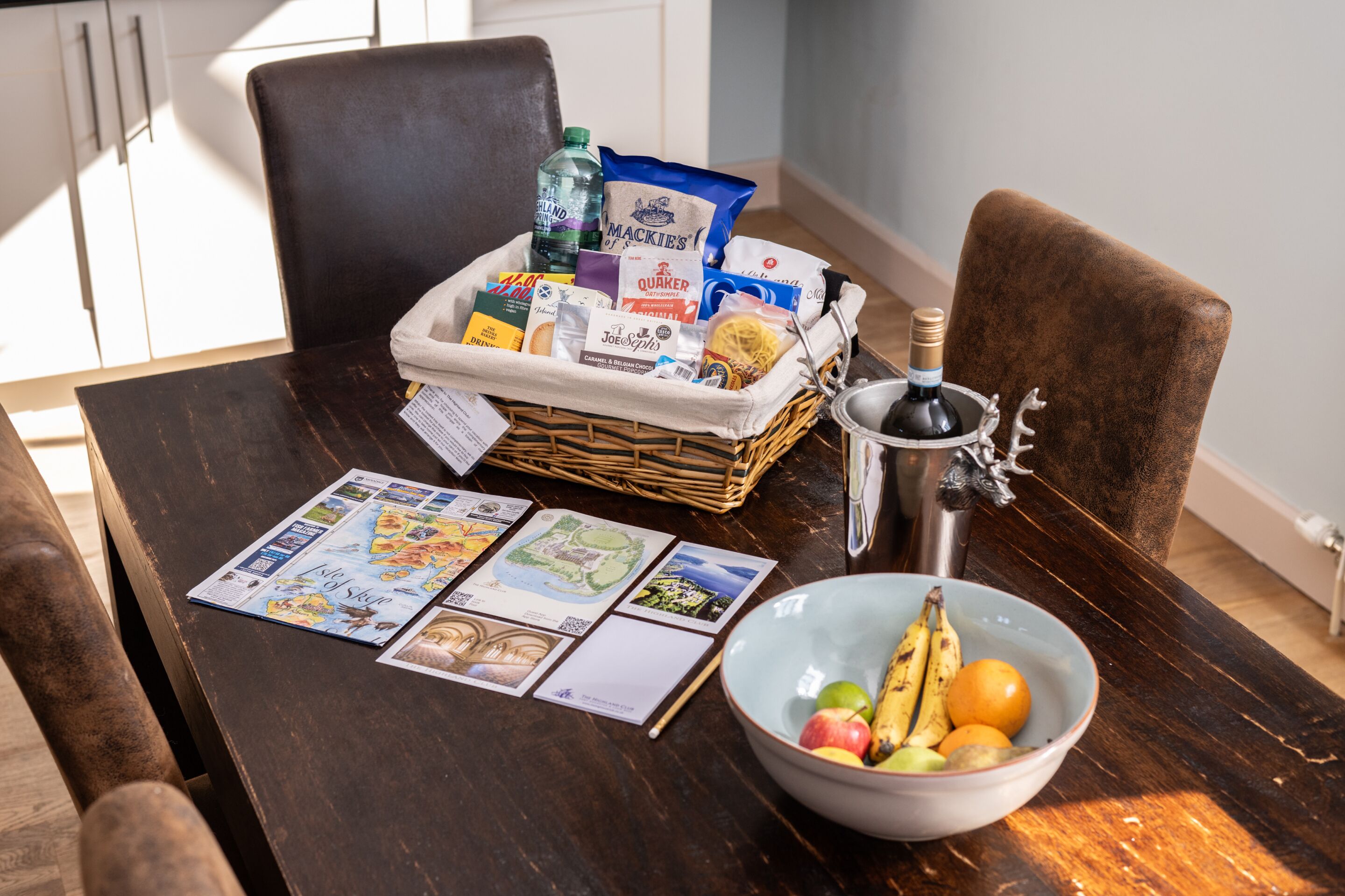 A dining table with a wicker basket containing snacks, a bottle of water, and a bottle of wine. A bowl of fruit is on the table, and there are menus and a wine glass.