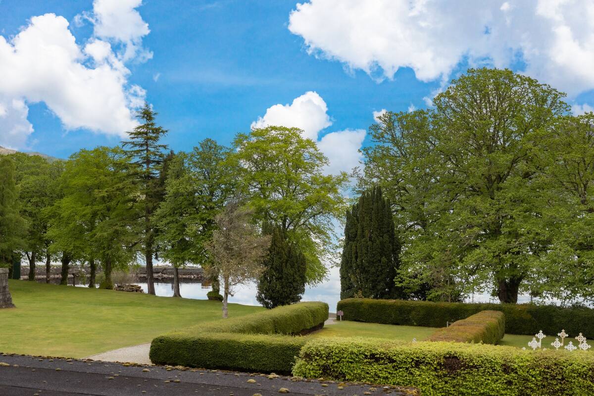 A well-maintained garden with neatly trimmed hedges, a variety of trees, and a calm body of water in the background.
