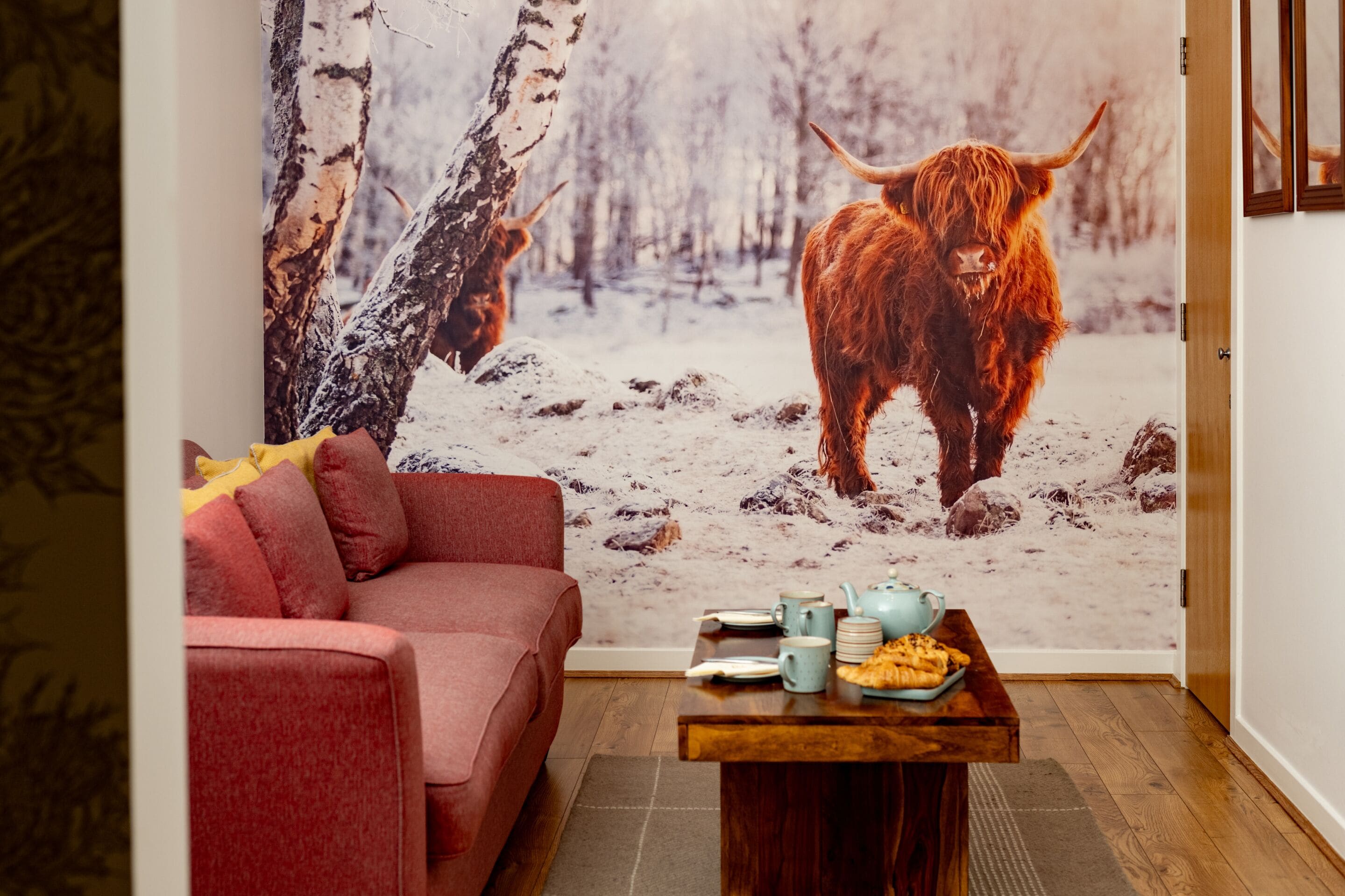 Una habitación acogedora con un sofá rojo, una mesa de centro de madera con una tetera y platos, y un mural grande en la pared que muestra un paisaje nevado con un yak.
