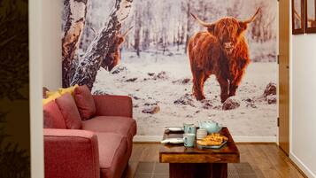 Una habitación acogedora con un sofá rojo, una mesa de centro de madera con una tetera y platos, y un mural grande en la pared que muestra un paisaje nevado con un yak.