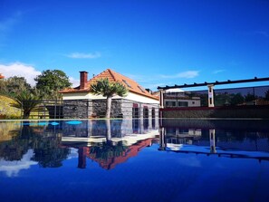 Piscina com reflexos nítidos, parede de pedra e edifício com telhado em azulejos.