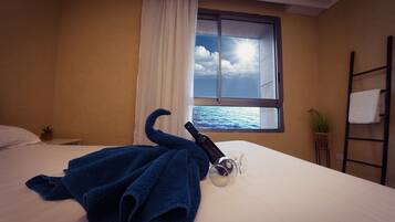 A hotel room with a bed, a window with a view of the sea, a towel rack, and a small plant.