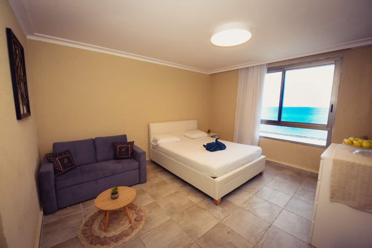 Quarto com cama, sofá, mesa redonda pequena e janela vista para o mar.
