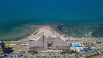 A large hotel with a central entrance, a parking lot, and a beach area with umbrellas.