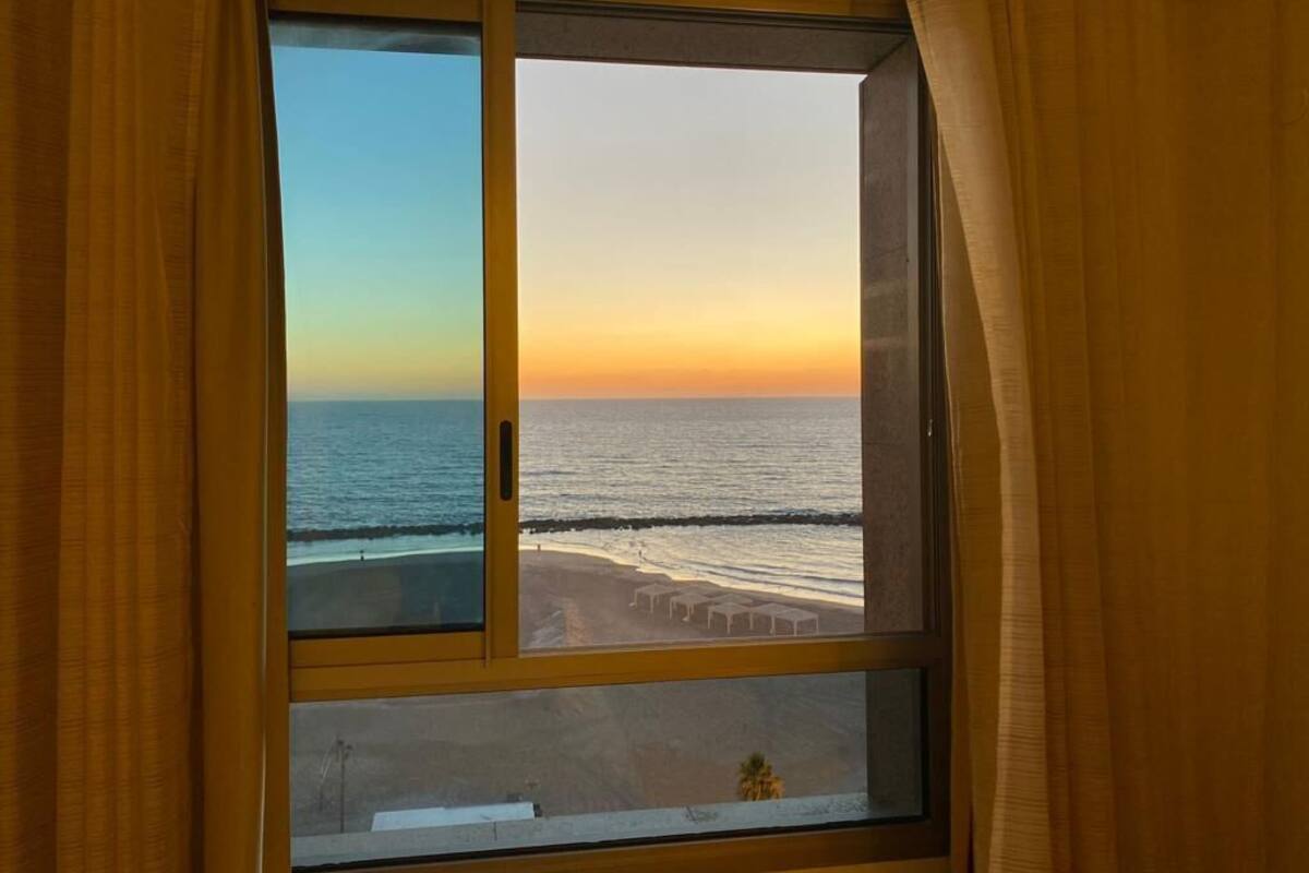 Quarto com uma janela ampla de frente para uma praia ao pôr do sol.