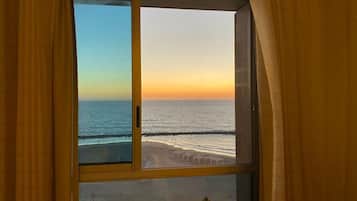 A room with a large window overlooking a beach at sunset.