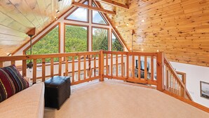 Ein Zimmer mit hohen Holzbalken, einem Balkon, großen Fenstern und Blick auf eine bewaldete Landschaft.