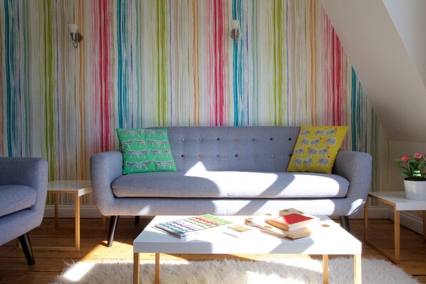 A modern living room with a striped wallpaper, a grey sofa with patterned cushions, a white coffee table with books and a plant, and a wooden floor. - Gorgeous, peaceful & very central apartment with fabulous views. (Bath)