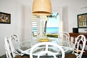 A dining area with a glass table, white chairs, a wicker pendant light, and a view of the ocean.