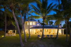 A modern house with a circular section, surrounded by palm trees and a well-lit outdoor area with seating.