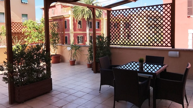 A covered outdoor area with tiled flooring, potted plants, wicker furniture, and a view of buildings in the background. - DELICIOUS LOFT TO SAN PIETRO (Roma)