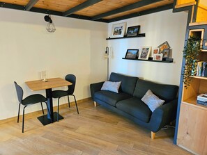 A cozy living area with a wooden table, two chairs, a dark sofa with patterned cushions, and a bookshelf with books and a plant.