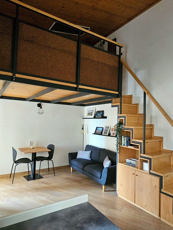 A modern interior with a wooden staircase, a sofa, a dining table with chairs, and a small bookshelf.
