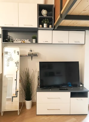 Una sala de estar moderna con un televisor de pantalla plana, muebles blancos, un estante con adornos y una planta en maceta.