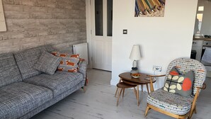 A living room with a grey patterned sofa, a round wooden table with a lamp, and a chair with a newspaper cushion. - Breathtaking Ocean Views,  Luxury Beachside Apartment, Freshwater Bay -  AONB (Freshwater Bay)