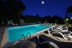 Una piscina con sillas de descanso y un tobogán, rodeada de árboles y una luna en el cielo.