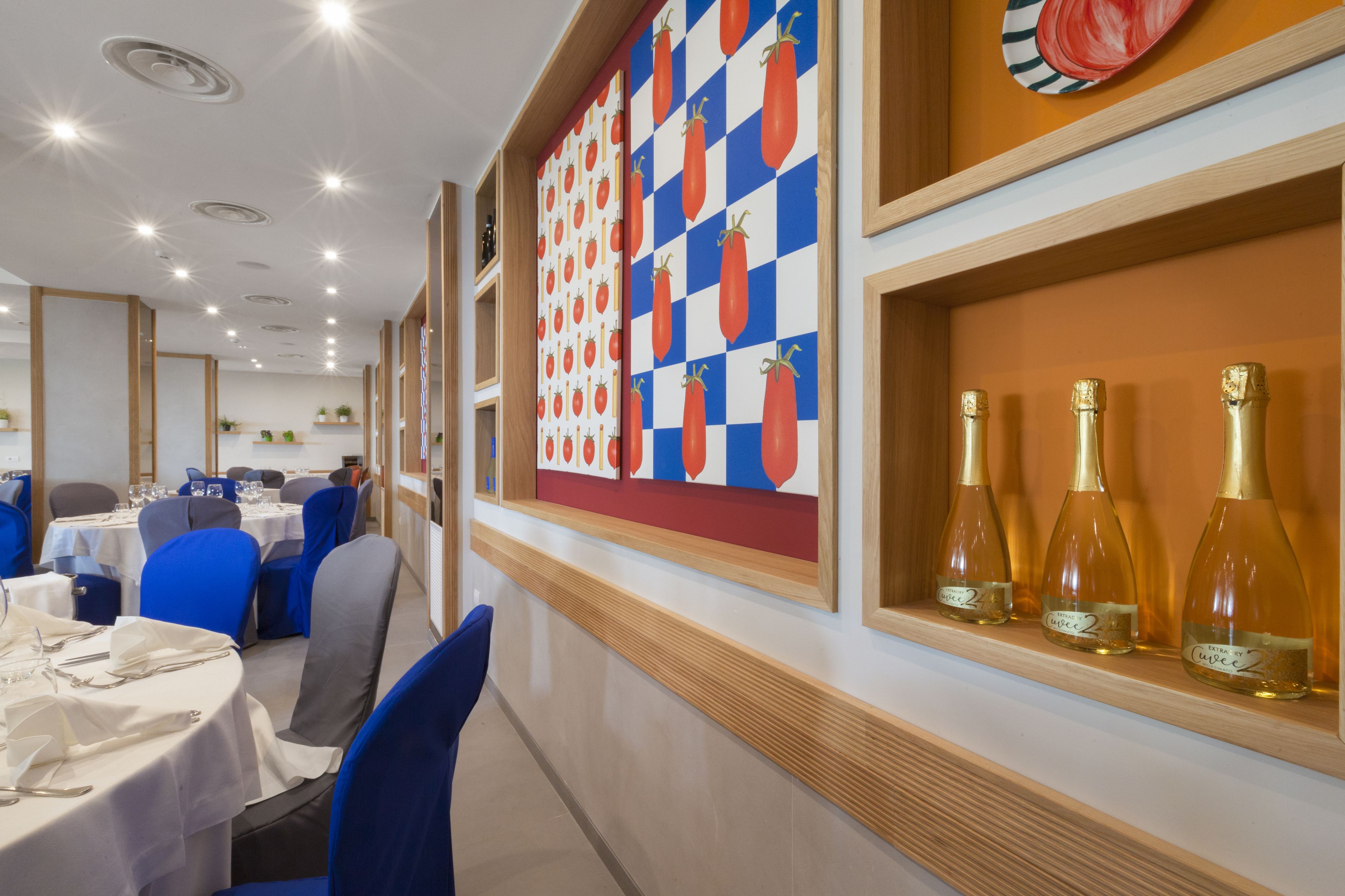 Un restaurant avec des tables dressées pour le service, des chaises bleues et un mur orné d’un motif de tomates.