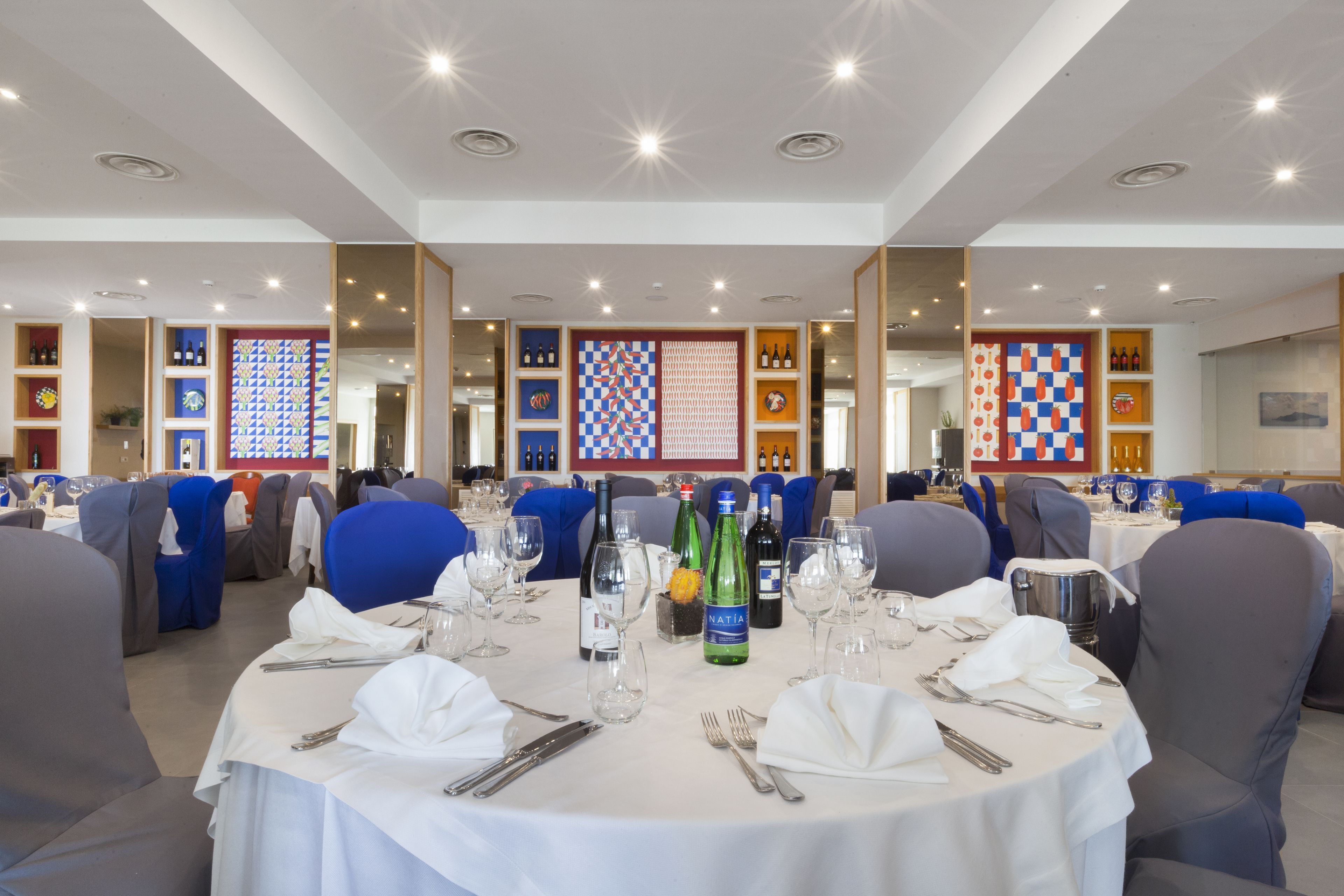 A dining area with round tables set for a meal, each with a white tablecloth, silverware, and wine bottles.