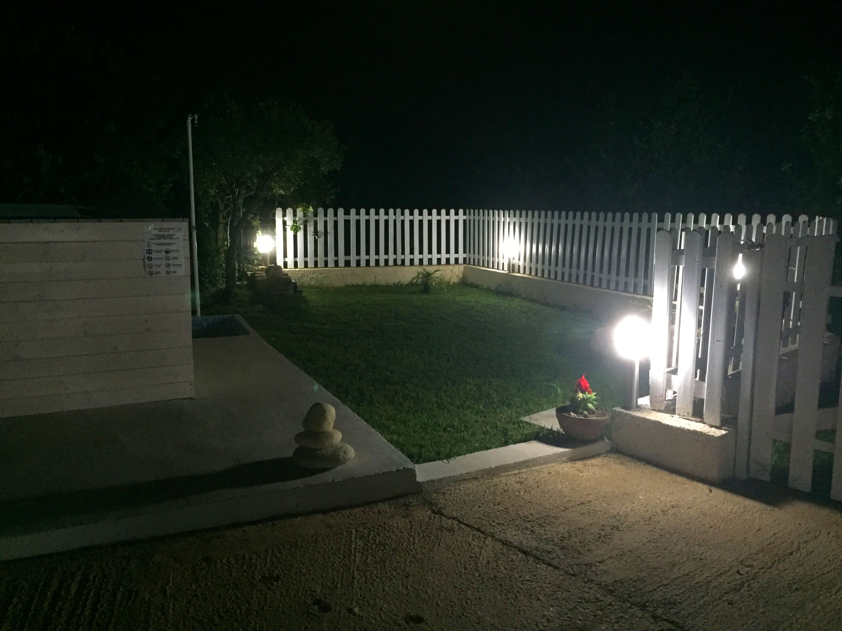 A gated entrance with a white picket fence, a small garden area with a potted plant, and a concrete pathway leading to the gate.