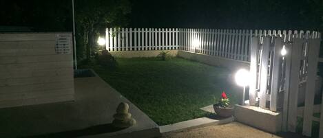 A gated entrance with a white picket fence, a small garden area with a potted plant, and a concrete pathway leading to the gate.