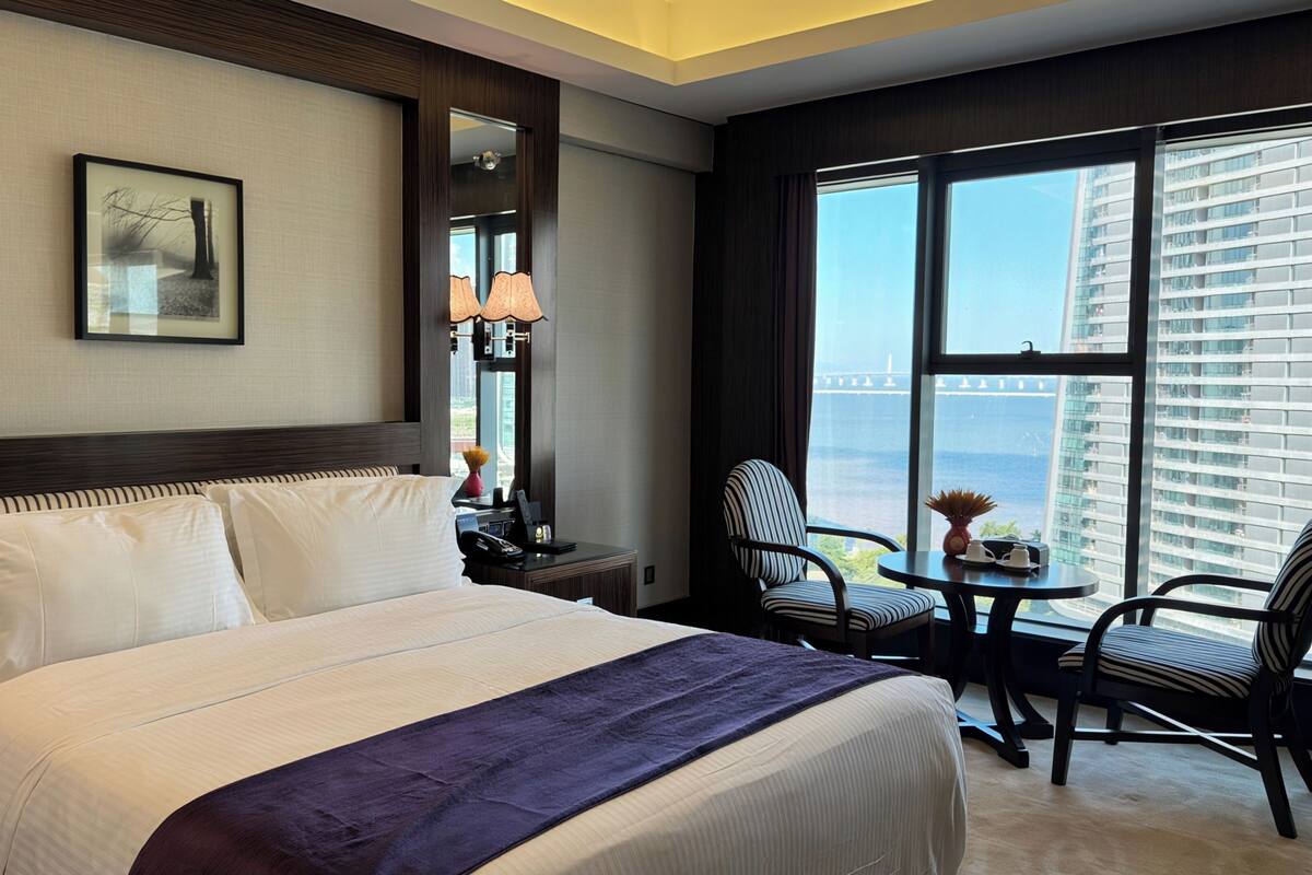 A hotel room with a large bed, a desk with a chair, a view of the cityscape, and a large window.