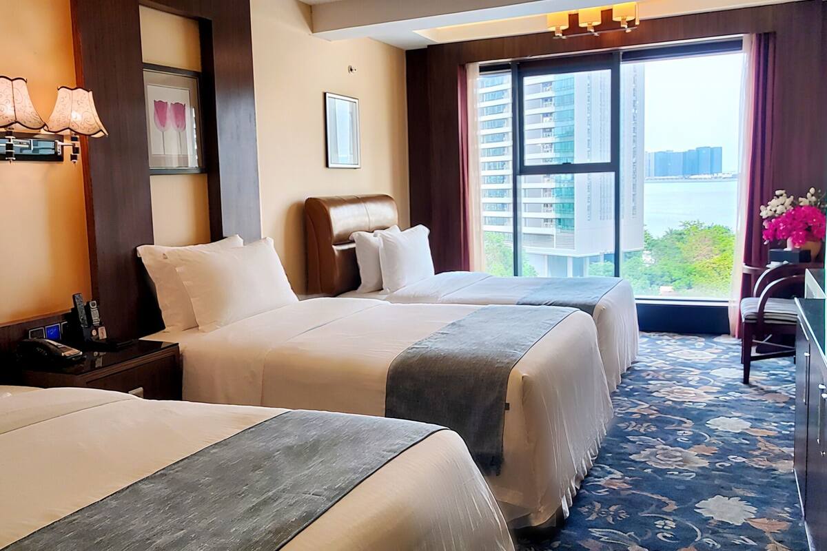 A hotel room with two beds, a desk, a chair, and a view of the city.