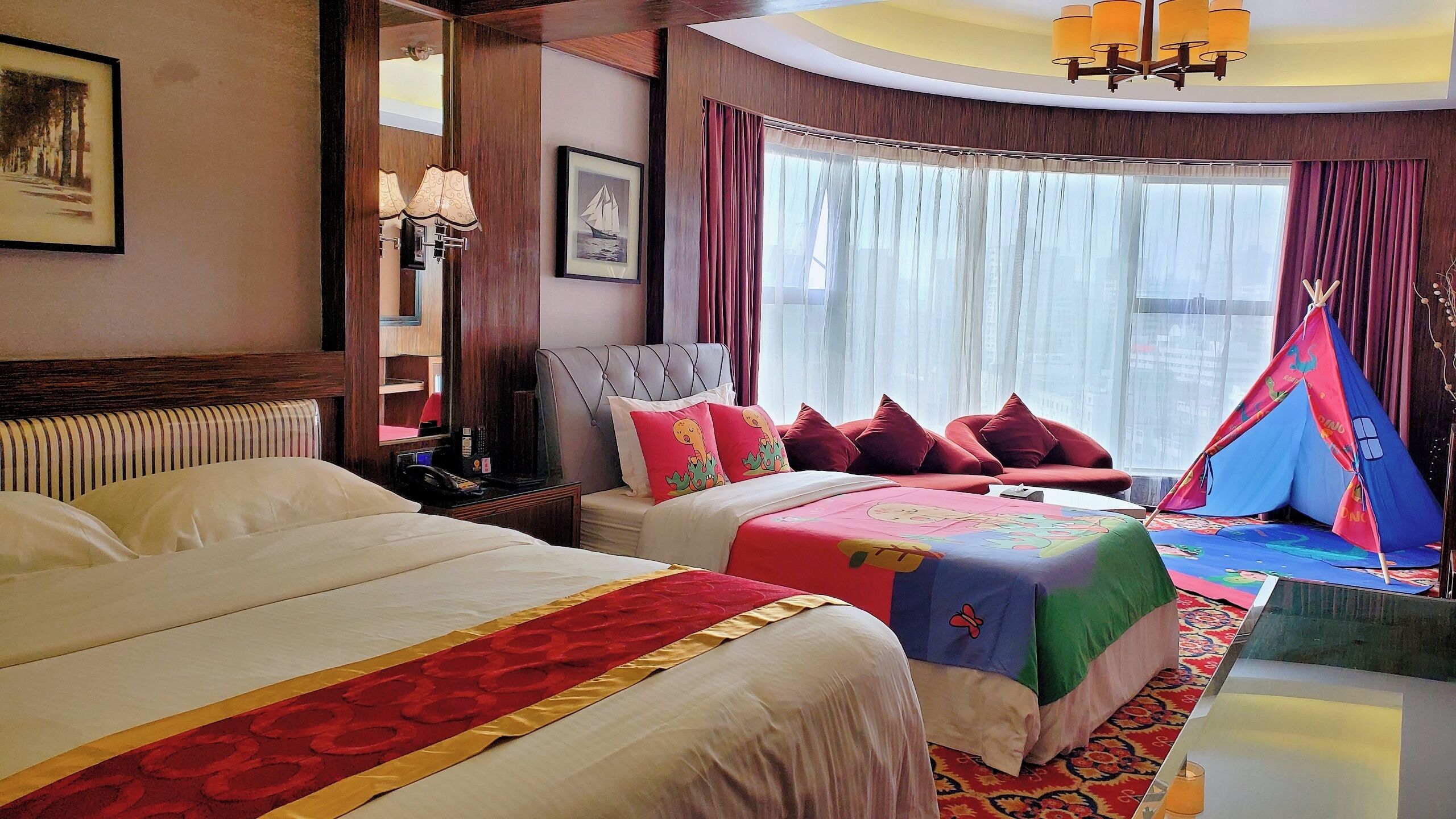 A hotel room with two beds, a window with sheer curtains, a red and gold patterned bed runner, a bedside table with a lamp, and a tent-like structure near the window.