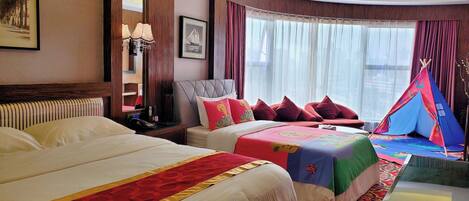 A hotel room with two beds, a window with sheer curtains, a red and gold patterned bed runner, a bedside table with a lamp, and a tent-like structure near the window.