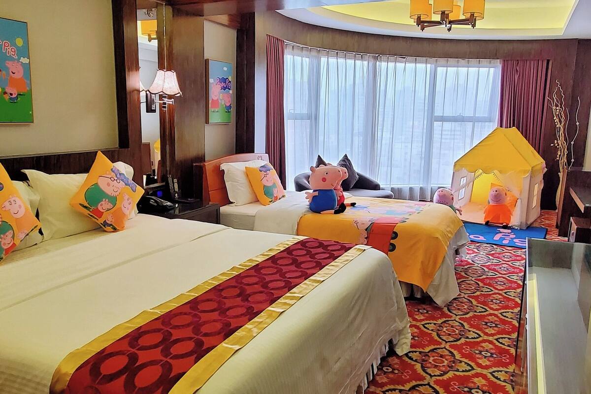 A hotel room with two beds, Peppa Pig-themed pillows, a large window with curtains, and a small Peppa Pig house.