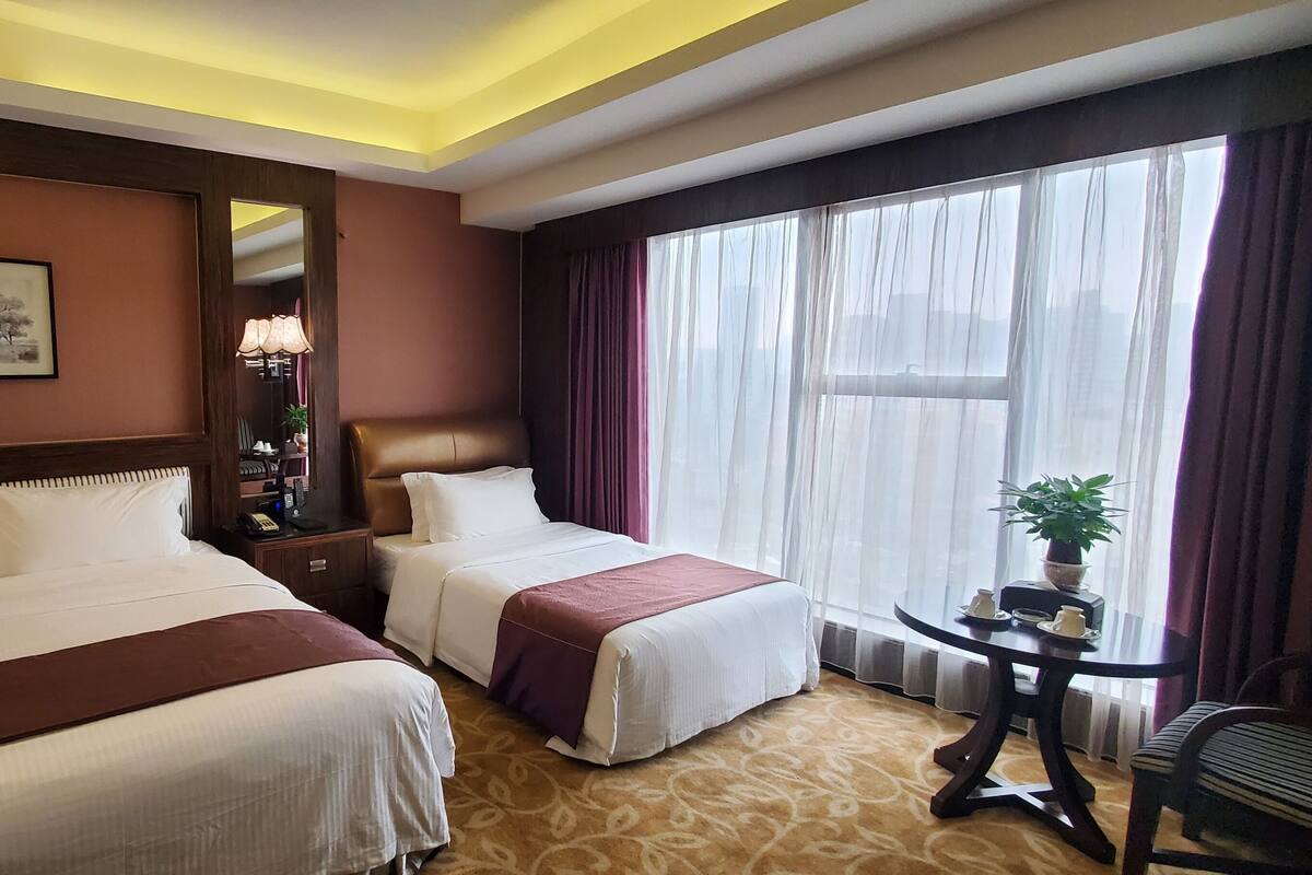 A hotel room with two beds, a large window with curtains, a small table with a plant, and a chair.