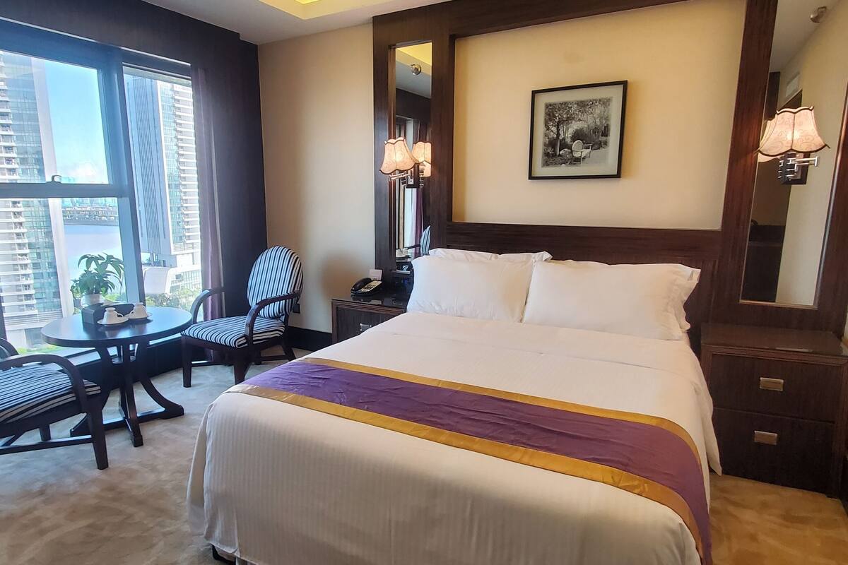 A hotel room with a large bed, a desk with a chair, a small table with a plant, and a large window offering a city view.