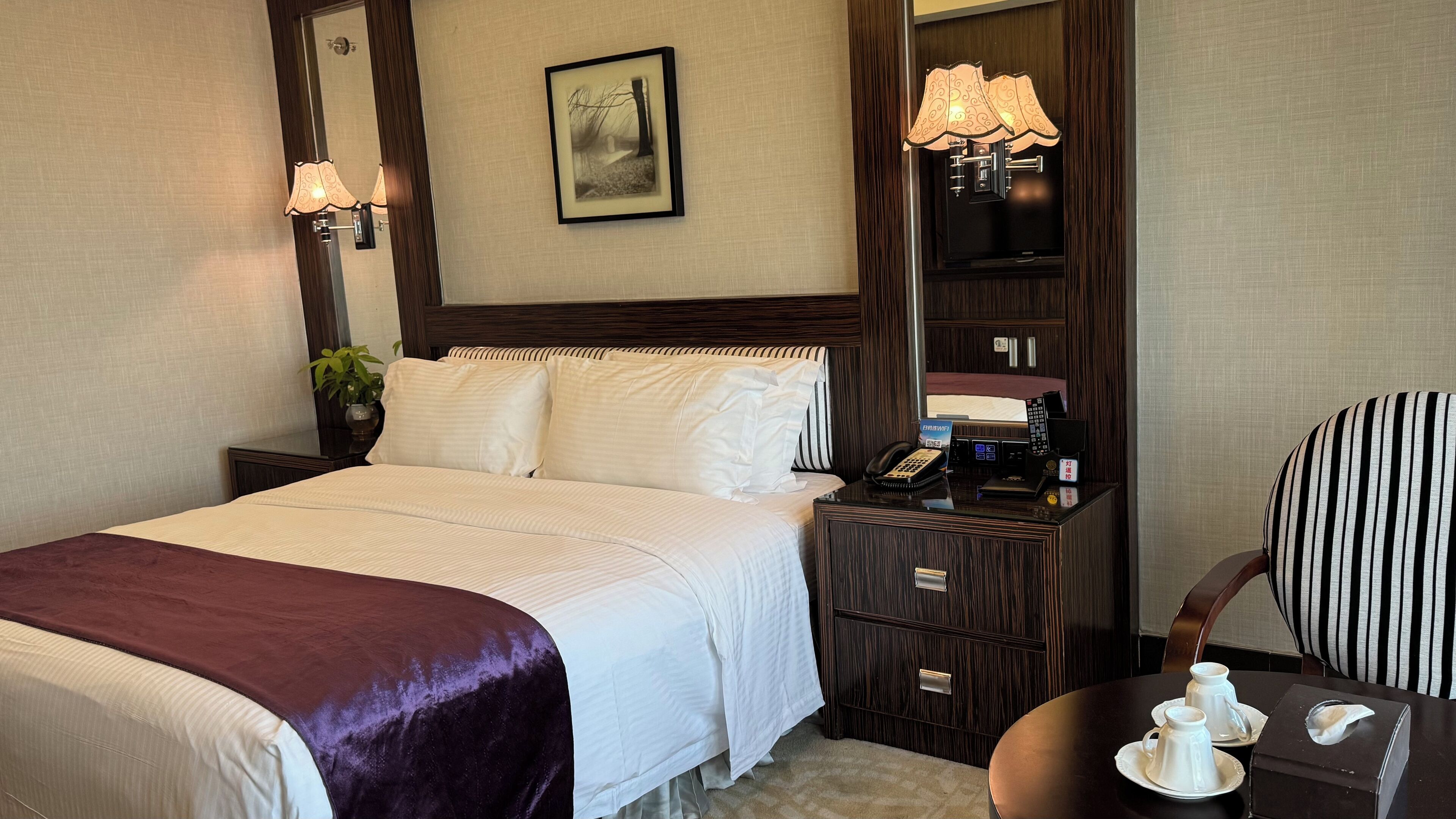 Une chambre d’hôtel avec un grand lit, une table de chevet avec un téléphone et une lampe, une chaise, et une petite table avec une théière et des tasses.
